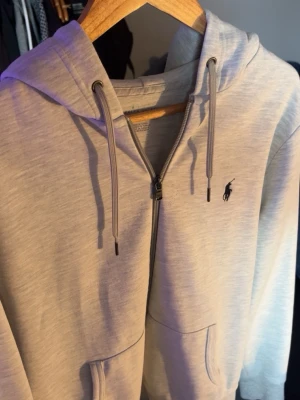 Ralph lauren zip - Säljer en stilren ljusgrå hoodie från Ralph Lauren med dragkedja och huva. Tröjan har snörning i huvan och den klassiska svarta Polo-loggan broderad på bröstet. Perfekt för en avslappnad look. Jätte fint skick och inga slitningar förutom att de är lite nopprigt vid bröstet men syns knappt, skriv privat om du har några funderingar!