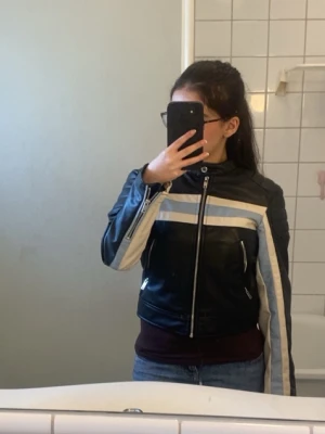 Stradivarius Svart skinnjacka med vita ränder - Snygg svart skinnjacka med vita och ljusblå ränder över bröstet och längs ärmarna. Jackan har ståkrage, dragkedja framtill och dragkedjor vid fickor och ärmslut. Perfekt för en cool och sportig look. Använd endast 2-3 gånger.