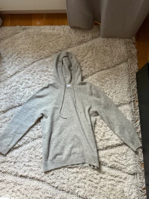 Ljusgrå Merin wool tröja  - Säljer en stilren ljusgrå hoodie med dragsko i huvan. Tröjan har långa ärmar, ribbade muddar och är tillverkad i en mjuk ullblandning som känns riktigt skön mot huden. Perfekt för dig som gillar en clean och avslappnad look.