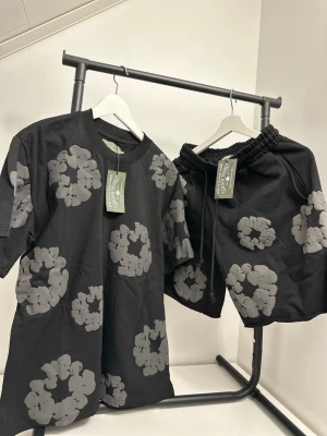 Svarta shorts med grått blommönster - Säljer ett par svarta shorts Denim tears med grått blommönster. Shortsens design är loose fit med elastisk midja och snörning. Materialet är mjuk bomull och de har en avslappnad look som passar perfekt till sommaren.