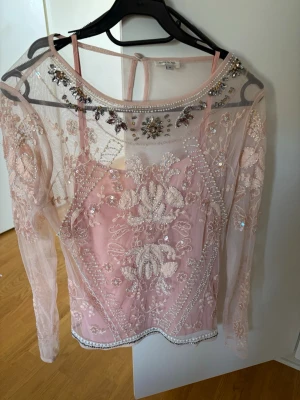Rosa blus med spets och pärlor - Säljer en romantisk rosa blus från Zara med transparent spets och broderade blommor. Blusen har långa ärmar och är dekorerad med pärlor och glittrande detaljer vid halsen. Perfekt för dig som gillar unika plagg med mycket detaljer.