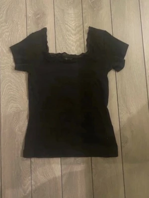 Svart topp med spets från Filippa K - Säljer en svart topp från Filippa K med snygga spetsdetaljer runt halsen. T-shirten har korta ärmar och en feminin, slim passform som framhäver siluetten. Perfekt att matcha med jeans eller kjol för en cool look.