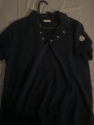 Mörkblå kortärmad skjorta från Moncler - Säljer en mörkblå skjorta från Moncler med klassisk krage och knappar framtill. Skjortan har korta ärmar och Moncler-loggan på ena ärmen. Perfekt för dig som vill ha en stilren och exklusiv look.