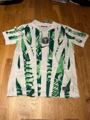 Nigeria fotbollströja Nike grön/vit - Säljer en Nigeria fotbollströja från Nike i vitt med gröna grafiska mönster. Tröjan har korta ärmar, rund hals och Nigeria-emblemet på bröstet. Tillverkad i lätt och ventilerande material, perfekt för träning eller match.