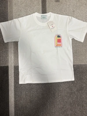 Vit t-shirt från Casablanca - Vit t-shirt från Casablanca med färggrafiskt tryck på bröstet och ryggen. Trycket föreställer en båge i rosa, gult, svart och rött med texten 'Casa Blanca'. T-shirten har rund hals och korta ärmar, tillverkad i mjuk bomull.