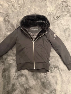 Svart hoodie från Moose Knuckles - Snygg svart hoddie/jacka från Moose Knuckles med dragkedja framtill och mjukt foder i huvan. Den köptes 1-2 år sen och har bara legat i garderoben, kvitto/bevis på att den är äkta finns såklart. Den är i storlek S men passar en medium då jag bär S men den är lite stor på mig. Jag har totalt använt den max 10-15 gånger då jag inte tyckte om den lika mycket som jag önskat