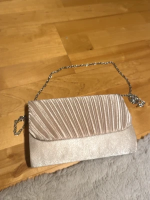 Silverfärgad kuvertväska med kedja - Stilren kuvertväska i skimrande silver med veckad framsida och elegant kedjerem. Perfekt accessoar för att lyfta din outfit. Väskan har en klassisk rektangulär form och stängs med lock. Materialet är glansigt tyg som ger en lyxig känsla.