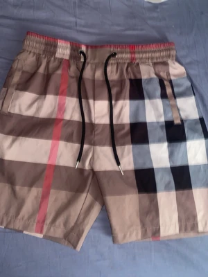 Rutig shorts från Burberry med snörning - Säljer ett par klassiska Burberry shorts med rutigt mönster i beige, svart, vit och röd. Shortsen har elastisk midja med svart snörning och sidofickor. Tillverkade i mjukt bomullsmaterial, perfekta för sommaren.Rutig shorts från Burberry med snörning