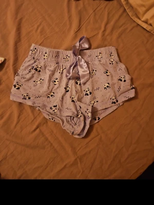 Lila pyjamasshorts med pandamönster - Söta lila pyjamasshorts med elastisk midja och stor satinrosett framtill. Shortsen har ett gulligt pandamönster och små 'zzz'-detaljer, perfekta för mysiga hemmakvällar. Tillverkade i mjukt och lätt material för extra komfort.