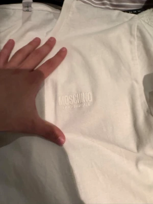 Vit Moschino t-shirt med logga - Säljer en vit t-shirt från Moschino med diskret broderad logga på bröstet. Klassisk passform och rund halsringning, perfekt för dig som gillar stilrena plagg med designer-touch. Materialet är mjukt och känns skönt mot huden.