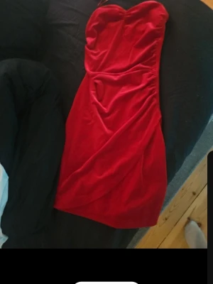 Röd axelbandslös sammetsklänning - Axelbandslös röd klänning i mjuk sammet med draperad effekt på sidan. Klänningen har en hjärtformad överdel och är kort, perfekt för dig som vill ha en elegant och trendig look. Passformen är tight och materialet känns lyxigt.