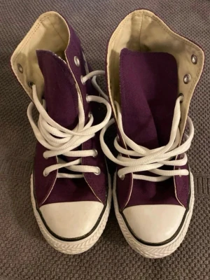 Lila Converse high-top sneakers - Säljer ett par lila Converse high-top sneakers med vita gummisulor och vita snören. Skorna har klassisk design med canvasmaterial och metallöglor. Insidan är ljusbeige och sulan har svart rand runt om. Perfekt för dig som vill ha en cool och färgstark look.