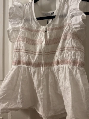 Vit blus med volang från RYVL S - Söt vit blus från RYVL S med volang och korta ärmar. Blusen har rosa broderade detaljer och knytning framtill. Tillverkad i ett lätt och luftigt material, perfekt för en avslappnad och trendig look.