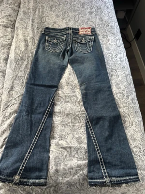 True Religion blå bootcut jeans - Säljer ett par blå bootcut jeans från True Religion med vita kontrastsömmar och snygga broderade bakfickor. Jeansen har slitningar och detaljer längs bensluten samt klassisk femficksdesign. Perfekt för dig som gillar en cool och avslappnad stil. Uppsprätta längst ner