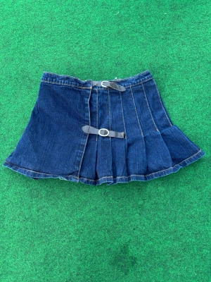 Mörkblå  jeanskjol med justerbar midja  - Säljer  en mörkblå plisserad jeanskjol med två dekorativa bälten framtill. Juster bar midja ✨ kolla bilden för måtten !!Kjolen har en kort och utställd modell med markerade sömmarf som ger en cool och edgy look. Perfekt för dig som vill sticka ut med en unik denimstil. PRIS KAN DISKUTERAS 