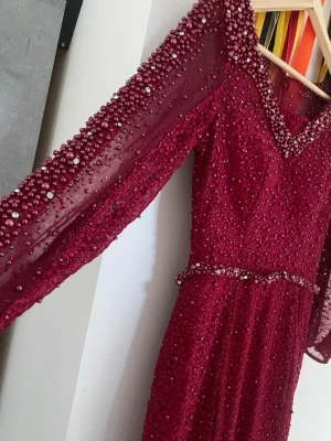 Vinröd långklänning med pärlor - Elegant vinröd långklänning med v-ringning och trekvartsärm. Klänningen är täckt av pärlor och har transparenta ärmar med extra dekorationer. Midjan markeras av ett pärlband och materialet ger en glittrig effekt. Perfekt för speciella tillfällen.använd 1 gång 