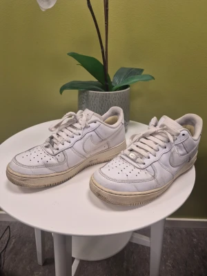 Nike Air Force 1 Vita Sneakers, Storlek 41 passar 42 - Säljer ett par klassiska vita Nike Air Force 1 sneakers med låg profil. Skorna har perforerad tå, snörning och den ikoniska swoosh-loggan på sidorna. Yttermaterialet är läder och sulan är i gummi. Perfekta för dig som gillar streetstyle och tidlös design.