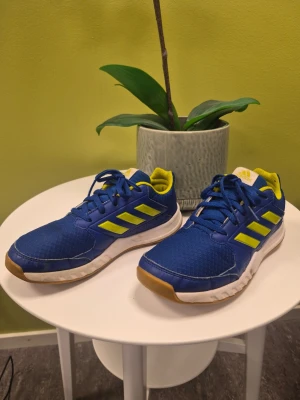 Adidas fortagym K Träningsskor blå/gul storlek 36 - Snygga blåa sneakers från Adidas med gula detaljer och klassiska tre ränder på sidan. Skorna har meshmaterial på ovansidan, vit sula med bruna inslag och en sportig design. Perfekta för dig som gillar en färgstark look och vill ha bekväma skor till vardags.