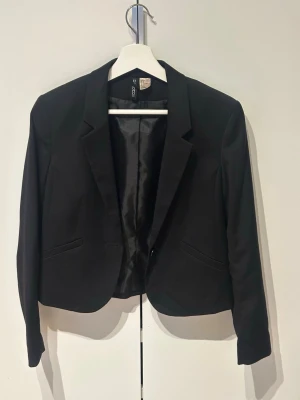 Svart kavaj från H&M med knapp - Stilren svart kavaj från H&M med klassisk krage och en knapp framtill. Kavajen har två fickor och är helfodrad med slätt tyg på insidan. Passformen är figurnära och modellen är kortare, perfekt för att skapa en snygg och modern look.