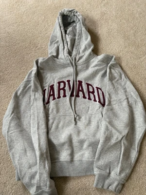 Grå hoodie - Snygg grå hoodie med stor huva och dragsko, samt magficka framtill. På bröstet står det 'HARVARD' i stora vinröda bokstäver. Tillverkad i mjukt bomullsmaterial, perfekt för en avslappnad och cool stil.