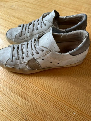 Vita Philippe Model sneakers - Snygga vita sneakers från Philippe Model med grå detaljer och klassisk låg siluett. Skorna har en ikonisk sidologga, vita skosnören och ovandel i läder. Perfekta för dig som gillar en stilren och avslappnad look.