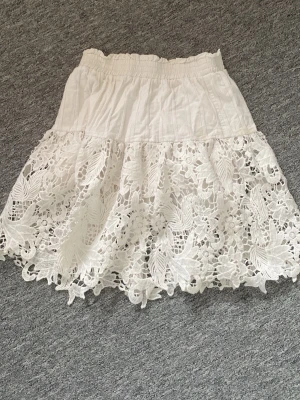 Kjol med spetsliknande blommor - Vit kjol med blommiga spetsdetaljet. Tyvärr har den några fläckar, plus lite nopprig.