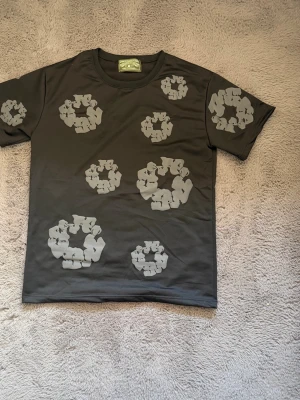 Mörkgrå t-shirt med grått mönster - Cool mörkgrå t-shirt från A Bathing Ape med unikt grått mönster som liknar blommor eller cirklar över hela tröjan. Klassisk passform och rund halsringning, perfekt för dig som vill sticka ut med en streetstyle-look.