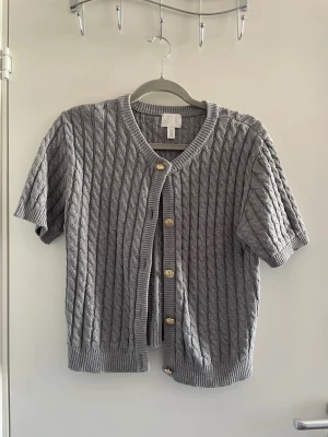 Grå stickad kofta med kort ärm - Säljer en grå stickad kofta från H&M med korta ärmar och snygga guldfärgade knappar framtill. Koftan har ett klassiskt kabelstickat mönster och rund halsringning. Perfekt att slänga över en t-shirt för en chill look.