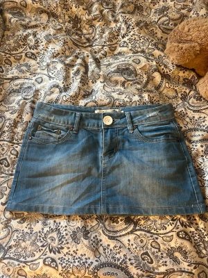 Blå jeanskjol med fickor - Säljer en blå jeanskjol med klassiska fickor fram och bak, vit knapp och hällor för bälte. Kjolen har en kort och rak passform, perfekt för dig som gillar denim. Snygga sömdetaljer och slitningar på bakfickorna ger en cool look.