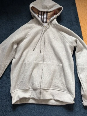 Grå hoodie med dragkedja från Burberry - Snygg grå hoodie från Burberry med dragkedja och klassiskt rutigt foder i huvan. Mjuk bomullsblandning, två stora fickor fram och justerbar dragsko i huvan. Perfekt för en chill och stilren look. Skriv för minsta lilla fundering