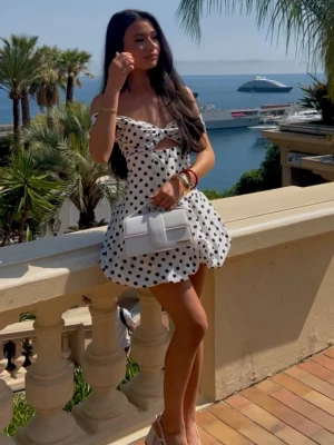 Vit prickig kort klänning med volang - Superfin vit kort klänning med svarta prickar och volang nertill. Klänningen har offshoulder-design, en liten cut-out vid bröstet och ärmlös stil. Perfekt för sommarens varma dagar och strandhäng. Materialet känns lätt och luftigt, troligen bomull eller polyester.