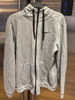 Grå zip-hoodie från H&M - Snygg grå hoodie med dragkedja från H&M. Modellen har huva med svarta snören, två stora fickor framtill och ribbade muddar. Perfekt för en avslappnad stil. Tillverkad i mjukt sweatshirtmaterial.