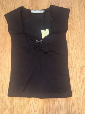 Svart topp med spetsdetalj från lager 157 - Svart ärmlös topp från 157 med v-ringning och spetsdetaljer längs ringningen. Toppen har en liten rosett framtill och är tillverkad i mjukt bomullsmaterial. 