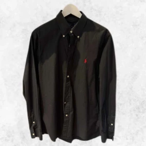 Svart skjorta från Ralph lauren - Snygg svart skjorta från Polo Ralph Lauren med röd broderad logga på bröstet. Skjortan har en slim fit passform och knappar framtill samt vid kragen. Perfekt för en stilren look.