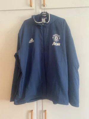 Blå Manchester United vindjacka Adidas - Blå vindjacka från Adidas med Manchester United-logga och Aon-tryck på bröstet. Jackan har huva, hel dragkedja och två sidofickor med dragkedja. Materialet är lätt och vindtätt, perfekt för att hålla sig skyddad mot blåst.
