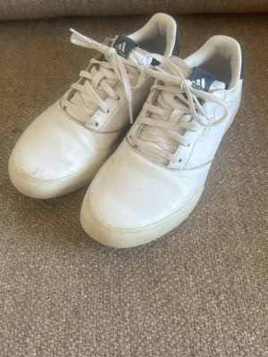 Golfskor - Vita Adidas - Säljer ett par vita sneakers/golfskor från Adidas med klassisk låg profil och snörning. Skorna har en stilren design med diskreta detaljer och är tillverkade i läder. Perfekta för golfen.