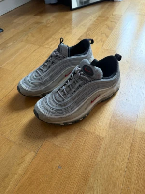 Nike Air Max 97 silver sneakers - Nike Air Max 97 sneakers i silver med röda detaljer och klassiska vågformade linjer. Ovandel i mesh och syntet, svart insida och synlig Air Max-sula. Ikonisk design med dragflik bak och framtill. Perfekta för dig som gillar streetstyle.