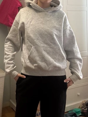 Grå basic hoodie med huva - En enkel och stilren grå hoodie med huva och stor magficka. Tröjan har ribbade muddar vid ärmslut och nederkant, och är gjord i mjukt sweatshirt-material. Perfekt för en avslappnad look och passar till det mesta.