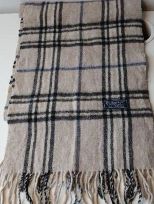 Beige rutig halsduk från Burberrys - Klassisk beige halsduk från Burberrys i 100% kashmir. Halsduken har ett rutigt mönster i svart, vitt och blått samt fransar i ändarna. Perfekt accessoar för att lyfta din outfit med en tidlös brittisk stil.