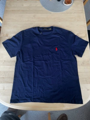 Mörkblå t-shirt från Polo Ralph Lauren - Säljer en mörkblå t-shirt från Polo Ralph Lauren med klassisk röd broderad logga på bröstet. T-shirten har rund hals och korta ärmar, perfekt för dig som gillar stilrena basics med en touch av ikonisk branding.