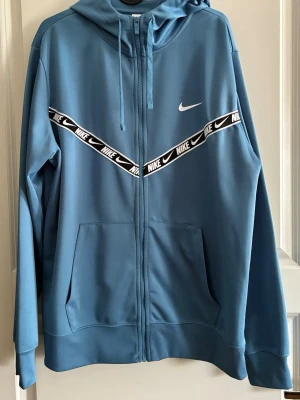 Blå Nike hoodie med dragkedja - Snygg blå hoodie från Nike med dragkedja framtill och huva med dragsko. Har en vit Nike-logga på bröstet och ett svartvitt band med Nike-tryck i V-form över bröstet. Praktiska fickor framtill och ribbade muddar vid ärmslut och nederkant.
