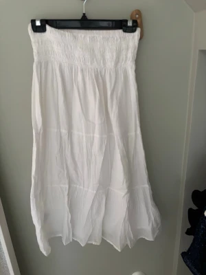 Vit långkjol från Gina Tricot - Säljer en vit långkjol från Gina Tricot med smock i midjan och lätt, luftig känsla. Kjolen har en flowy passform och är perfekt för varma dagar. Enkel och stilren design som passar till många olika looks.
