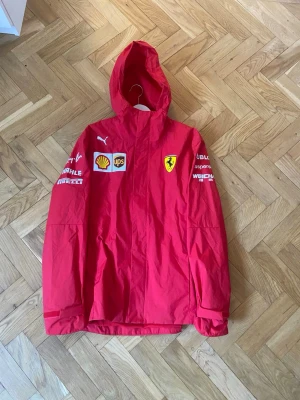 Röd Ferrari vindjacka från Puma - Snygg röd vindjacka från Puma i samarbete med Ferrari. Jackan har huva, dragkedja och flera loggor som Ferrari, Shell, UPS och Pirelli på bröstet och ärmarna. Tillverkad i lätt och tåligt material med meshfoder på insidan. Perfekt för dig som gillar motorsport och streetwear.