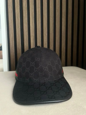 Svart Gucci keps med GG-mönster - Snygg svart keps från Gucci med klassiskt GG-mönster över hela kepsen. På sidan finns en detalj med gröna och röda ränder. Kepsen har böjd skärm och är tillverkad i canvasmaterial. Perfekt för dig som vill ha en exklusiv och trendig look.