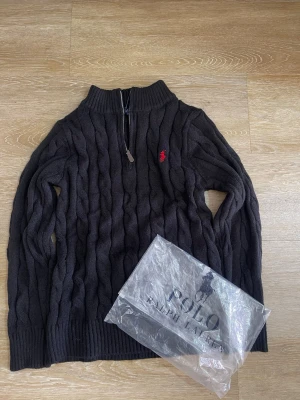 Mörkgrå kabelstickad tröja Polo - Mörkgrå kabelstickad tröja från Polo Ralph Lauren med halv dragkedja och ribbad krage. Tröjan har ett rött broderat logomärke på bröstet och är tillverkad i mjukt stickat material. Perfekt för lager-på-lager och snygg till jeans.