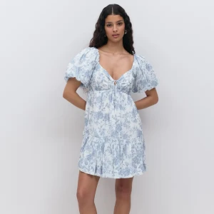 Blommig kortklänning från Gina Tricot - Supersöt kortklänning med blått blommönster på vit botten, puffiga kortärmar och v-ringning med snörning framtill. Klänningen har volang nertill, perfekt för varma dagar. Klänningen är använd en gång! Skriv för egna bilder💕