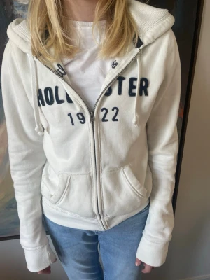 Vit hoodie från Hollister med tryck - Vit zip-hoodie från Hollister med mörkblå broderat tryck 'Hollister 1922'. Storlek M men sitter mer som S. Går att pruta! Skicka frågor om ni har någon.🥰