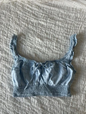 Ljusblå croppad linnetopp med knyt - Supersöt ljusblå croppad linnetopp med smockdetaljer och volangkantade axelband. Toppen har en liten rosett framtill och är perfekt för varma dagar. Materialet känns lätt och luftigt.