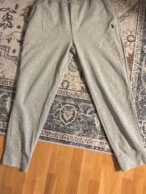 Grå mjukisbyxor från Polo Ralph Lauren - Snygga grå mjukisbyxor från Polo Ralph Lauren med elastisk midja och dragsko. Byxorna har två sidofickor och den klassiska svarta Polo-loggan broderad på vänster sida. Tillverkade i mjukt och bekvämt material, perfekta för chill dagar.