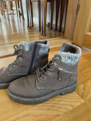 Grå stövletter med stickad kant - Snygga grå stövletter med grov sula och snörning framtill. De har en stickad mudd upptill i gråmelerat och dragkedja på sidan för enkel påtagning. Materialet ser ut att vara mockaimitation och skon har dekorativ liten ficka med dragkedja på utsidan.